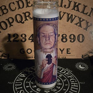 Puede incluir: Una vela blanca en un recipiente de vidrio transparente. La vela tiene una imagen impresa de un hombre con una expresión seria, sosteniendo un objeto oscuro. La imagen está rodeada por un borde azul con un patrón repetido de cruces. La vela está sentada sobre una superficie negra con un diseño de círculo blanco.