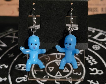 Op de afbeelding: Een paar blauwe babypop oorbellen met zilveren hoepels. De oorbellen worden getoond op een zwarte achtergrond met een witte cirkel en symbolen.