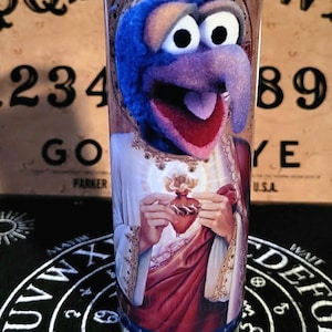 Może przedstawiać: Szklana świeca z niebieską i fioletową pluszową postacią trzymającą czerwone serce. Postać ma na sobie białą i złotą szatę z czerwoną przepaską. Świeca stoi na czarnej powierzchni z białym kołem i tekstem.