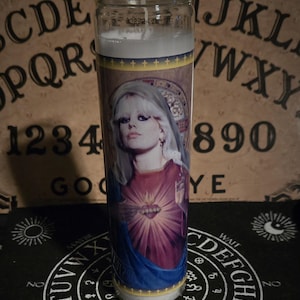 Puede incluir: Una vela blanca con una imagen colorida de una mujer con cabello rubio que lleva una túnica azul y morada. La imagen está rodeada por un marco dorado. La vela está sentada sobre una superficie negra con una tabla de Ouija blanca.