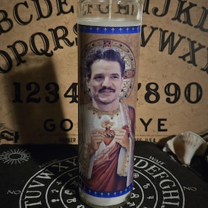May include: A glass candle with a portrait of a man in religious attire. The candle has a blue and gold band at the top and bottom. The background includes a Ouija board and a black mat with white lettering.