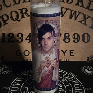 Puede incluir: Una vela blanca con una imagen impresa de una persona con cabello negro y una camisa negra sosteniendo un corazón. La vela está sobre una superficie negra con un círculo blanco y la palabra "YES" en el centro. Las palabras "OUIJA MYSTIFYING ORACLE" son visibles en el fondo.