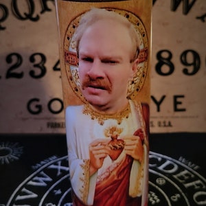 Może przedstawiać: Szklana świeca z religijnym wizerunkiem mężczyzny w białej szacie i czerwonych akcentach, trzymającego serce. Świeca stoi na czarnej powierzchni z białymi literami i symbolami. W tle znajduje się tablica Ouija.