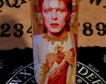 Puede incluir: Una vela blanca con una imagen de David Bowie en rojo, blanco y dorado en la parte delantera. La imagen muestra a Bowie con el pelo rojo, la cara blanca y un corazón de oro en las manos. La vela está sobre una superficie negra con un círculo blanco y las palabras "YES" y "NOW" impresas en ella.