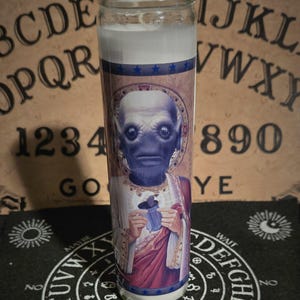 May include: A white candle with a blue and gold label featuring a portrait of a blue-skinned alien holding a cup. The candle is sitting on a black Ouija board with the letters "YES" and "NO" visible.