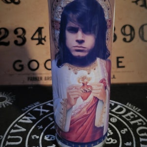 May include: A white glass candle with a printed image of a person with long dark hair holding a red heart. The person is wearing a white and red robe. The candle is sitting on a black surface with a white circle design.