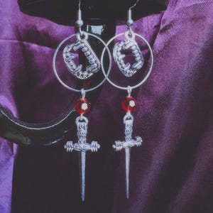 May include: Silver-toned dangle earrings with a gothic theme. Each earring features a silver hoop with a vampire fang charm, a red bead, and a small sword pendant. The earrings are displayed against a purple background.