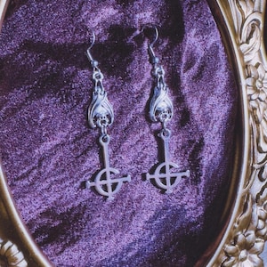 May include: A pair of silver-toned earrings. Each earring features a bat-like charm at the top, connected to a cross pendant. The earrings are displayed against a purple velvet background, framed by a gold-colored ornate border.