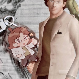 May include: A keychain featuring a cartoon character wearing a white lab coat and holding a book. The character has brown hair, glasses, and a smiling face. The keychain is attached to a white metal ring. The character is standing next to a person wearing a beige jacket and a green shirt. A green parrot is perched on the person's shoulder.