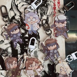 May include: A set of eight colourful character keychains featuring a variety of characters from the Metal Gear Solid video game series. The keychains are made of clear acrylic and feature detailed artwork of the characters. The keychains are attached to silver metal rings.