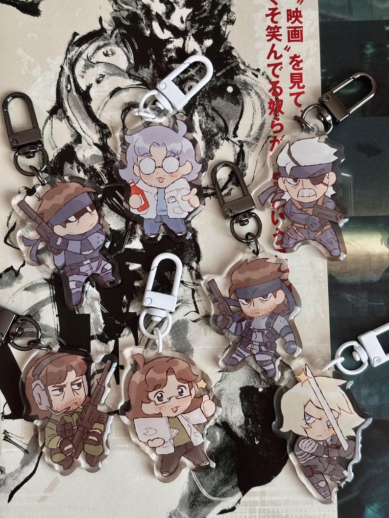 May include: A set of eight colourful character keychains featuring a variety of characters from the Metal Gear Solid video game series. The keychains are made of clear acrylic and feature detailed artwork of the characters. The keychains are attached to silver metal rings.