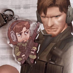 May include: A clear acrylic keychain featuring a cartoon character holding a rifle. The character has brown hair, a beard, and is wearing a green and brown military uniform. The keychain is attached to a silver metal ring.