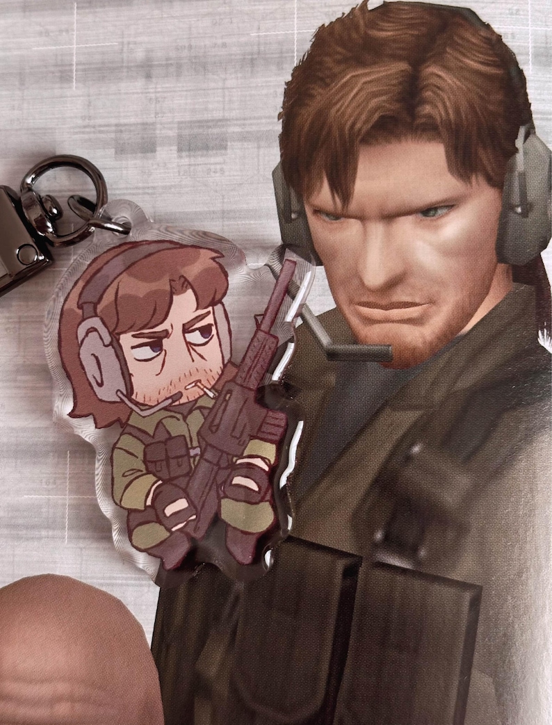 May include: A clear acrylic keychain featuring a cartoon character holding a rifle. The character has brown hair, a beard, and is wearing a green and brown military uniform. The keychain is attached to a silver metal ring.