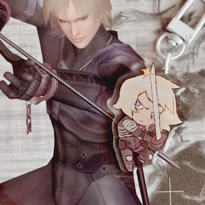 May include: A white keychain with a silver clasp and a cartoon character charm. The character is a blonde woman with a sword, wearing a grey and white outfit. The charm is attached to a black and silver chain.