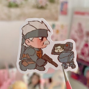 May include: A sticker featuring a cartoon illustration of a character with gray hair and a black eye patch, wearing a gray and brown outfit, holding a gun. The character is looking to the right at a small, gray robot with a screen for a head. The robot is wearing glasses and has a red light on its head.