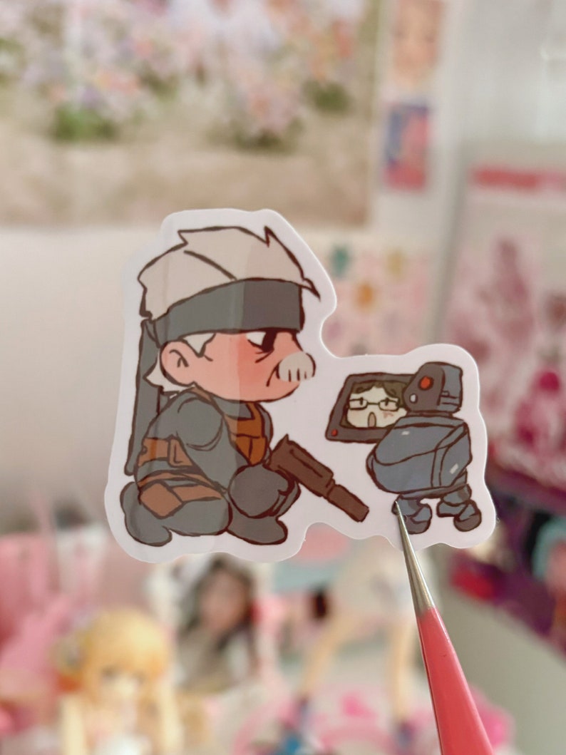 May include: A sticker featuring a cartoon illustration of a character with gray hair and a black eye patch, wearing a gray and brown outfit, holding a gun. The character is looking to the right at a small, gray robot with a screen for a head. The robot is wearing glasses and has a red light on its head.
