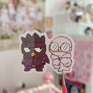 May include: Two cartoon stickers, one black and one white. The black sticker is of a bird wearing a purple ninja outfit and a black mask. The white sticker is of a white bird wearing a white coat and glasses.