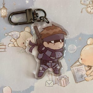 May include: A clear acrylic keychain featuring a cartoon character with brown hair, a purple bandana, and a black and grey outfit. The character is holding a gun and is standing on a white background.