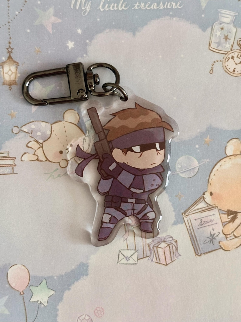 May include: A clear acrylic keychain featuring a cartoon character with brown hair, a purple bandana, and a black and grey outfit. The character is holding a gun and is standing on a white background.