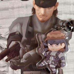 May include: A clear plastic keychain featuring a cartoon character holding a gun. The character is wearing a black eye patch, a black and white striped shirt, and a black belt with a silver buckle. The keychain is attached to a silver metal ring.