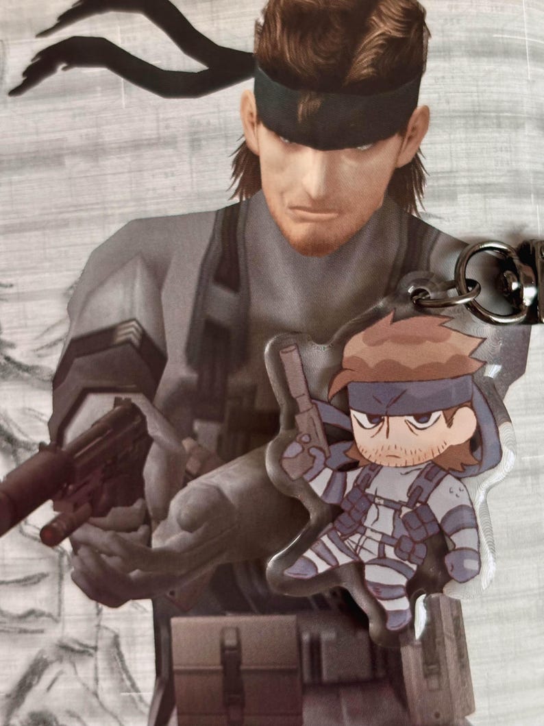 May include: A clear plastic keychain featuring a cartoon character holding a gun. The character is wearing a black eye patch, a black and white striped shirt, and a black belt with a silver buckle. The keychain is attached to a silver metal ring.