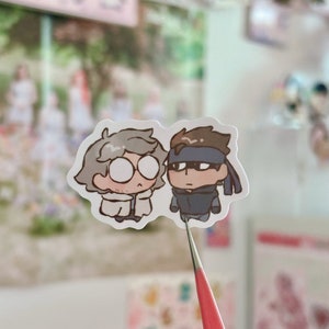 May include: Two cartoon characters, one with gray hair and glasses wearing a white coat and the other with brown hair wearing a blue jacket and a headband, are depicted on a white sticker.