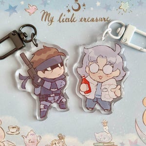 May include: Two anime-style character keychains. The keychain on the left features a character with brown hair, a black eye patch, and a gun. The keychain on the right features a character with white hair, glasses, and a red bottle. The text "My little treasure" is visible in the background.