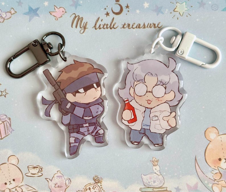 May include: Two anime-style character keychains. The keychain on the left features a character with brown hair, a black eye patch, and a gun. The keychain on the right features a character with white hair, glasses, and a red bottle. The text "My little treasure" is visible in the background.