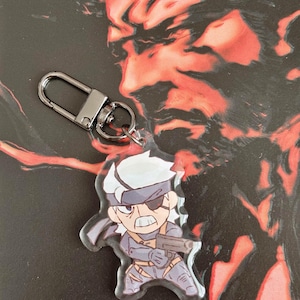 May include: A silver keychain with a clear acrylic charm featuring a cartoon character with grey hair, a black eyepatch, and a gun. The character is wearing a grey and black outfit.