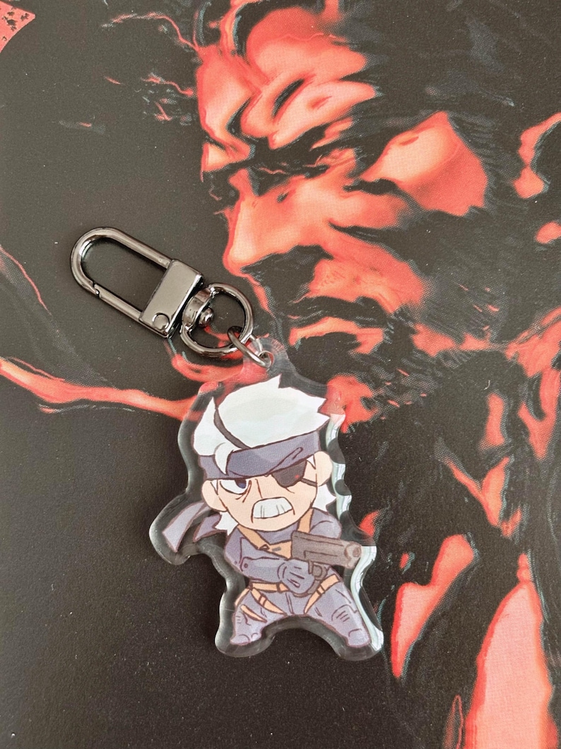 May include: A silver keychain with a clear acrylic charm featuring a cartoon character with grey hair, a black eyepatch, and a gun. The character is wearing a grey and black outfit.