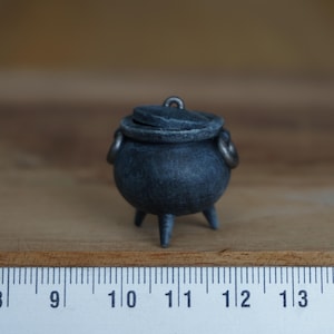 Könnte beinhalten: Ein kleiner schwarzer Kessel mit Deckel und drei Beinen. Der Kessel ist etwa 7,5 cm hoch und aus Metall.