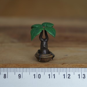 Op de afbeelding: Een klein, handgeschilderd beeldje van een boomwezen met een groene, bladerrijke top en een bruine, knoestige stam. Het wezen heeft een gezicht met een fronsende uitdrukking en zit in een bruine pot.