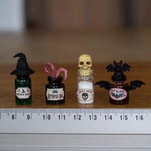 Peut inclure: Petites fioles de potions miniatures avec les étiquettes "Witch's Brew", "Octopus Ink", "Skull Dust" et "Bat Blood". Chaque flacon est surmonté d'un élément unique : un chapeau de sorcière, des tentacules, un crâne et des ailes de chauve-souris. Sur une surface en bois.