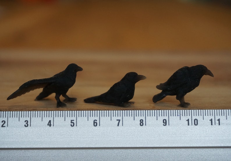 May include: Three black crow figurines, each about 2 inches long, are lined up on a wooden surface with a ruler for scale.