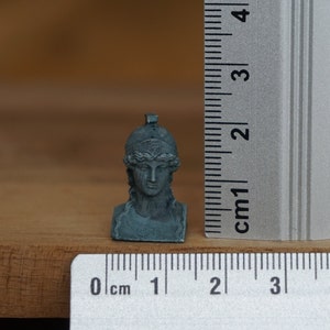 May include: A small, gray, sculpted bust of a woman wearing a helmet. The bust is approximately 2 cm tall and is sitting on a wooden surface.