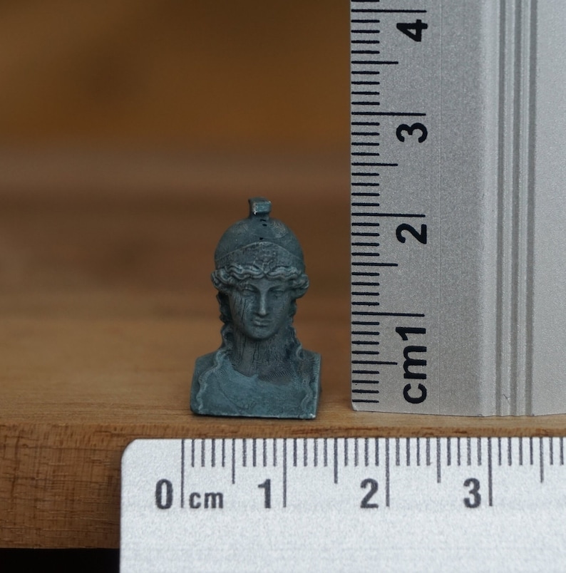 May include: A small, gray, sculpted bust of a woman wearing a helmet. The bust is approximately 2 cm tall and is sitting on a wooden surface.