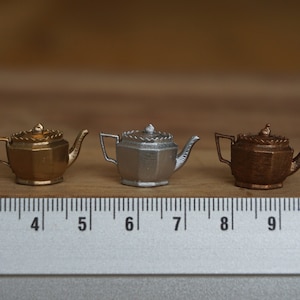 May include: Three miniature teapots, one gold, one silver, and one copper, are lined up on a wooden surface with a ruler underneath. The teapots are all the same design, with a rounded body and a spout on the side.