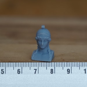 May include: A small, gray, 3D printed bust of a woman wearing a helmet. The bust is about 2 inches tall and is sitting on a wooden surface next to a ruler.