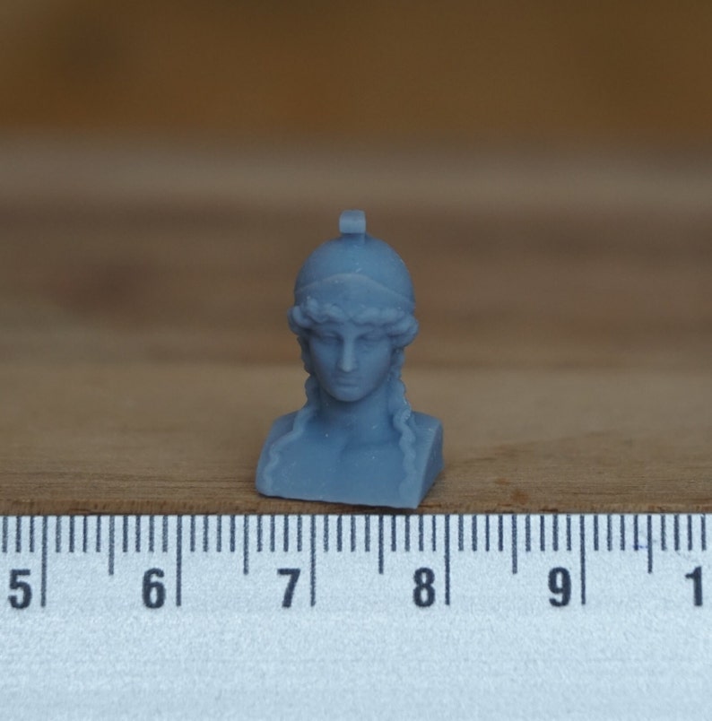 May include: A small, gray, 3D printed bust of a woman wearing a helmet. The bust is about 2 inches tall and is sitting on a wooden surface next to a ruler.