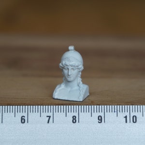 May include: White plaster bust of a woman wearing a helmet. The bust is about 2 inches tall and is sitting on a ruler.