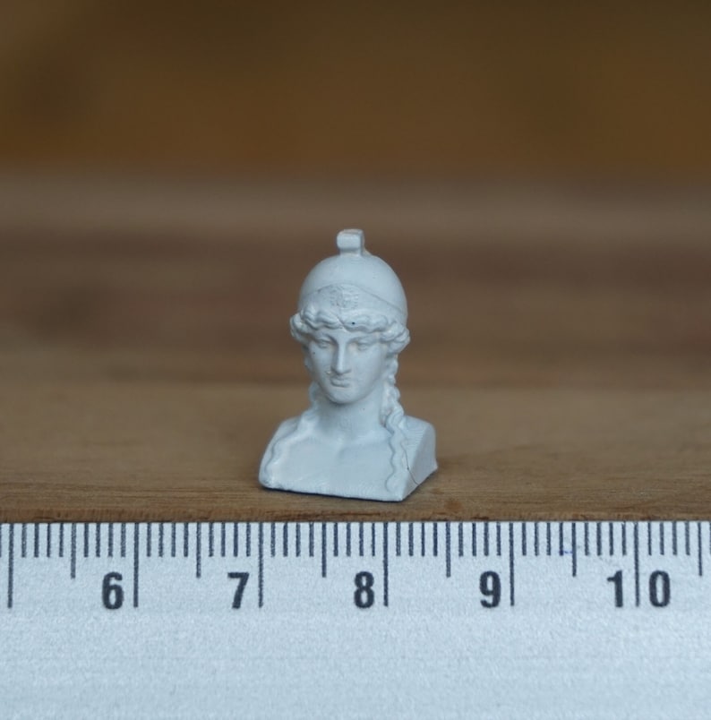 May include: White plaster bust of a woman wearing a helmet. The bust is about 2 inches tall and is sitting on a ruler.