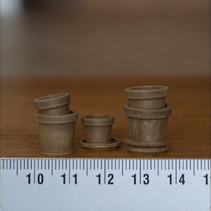 Op de afbeelding: Een verzameling kleine, bruine keramische bloempotten in verschillende maten. De potten hebben een klassiek ontwerp met een afgeronde vorm en een licht uitlopende rand. Een liniaal is eronder geplaatst voor de schaal.