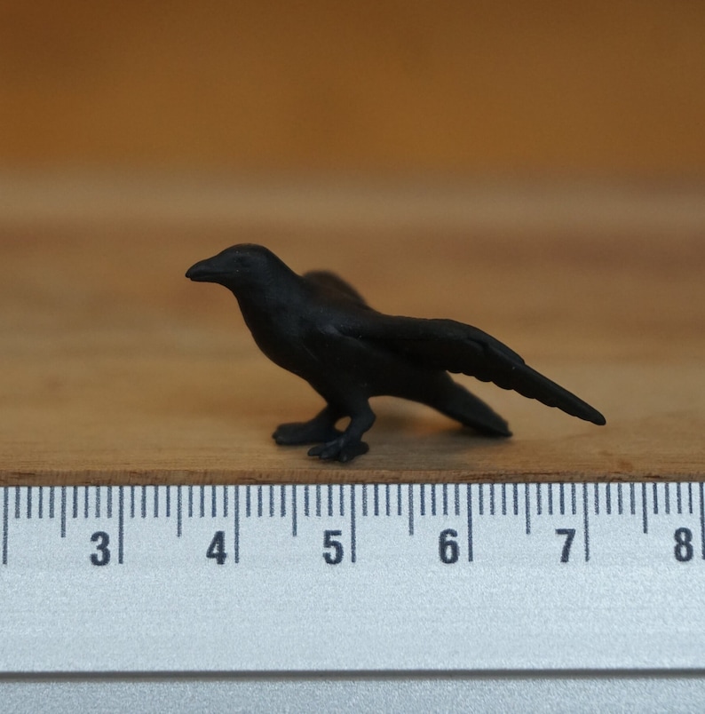 May include: A black crow figurine, approximately 5 inches long, is positioned on a white ruler with black markings. The crow is facing to the right with its wings slightly spread.