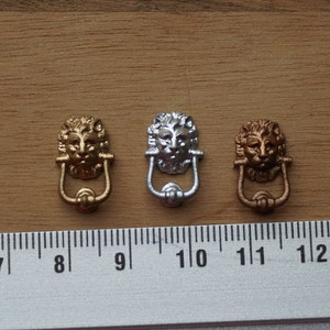 以下が含まれることがあります： 金、銀、銅の仕上げのミニチュアライオンヘッドドアノッカー3つ。ノッカーは約2.5cmの高さで、スケール用の定規が付いた木製の表面に表示されています。