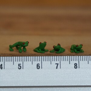Puede incluir: Cuatro pequeñas figuritas de rana verde con ojos rojos, alineadas en una regla con marcas del 4 al 8.
