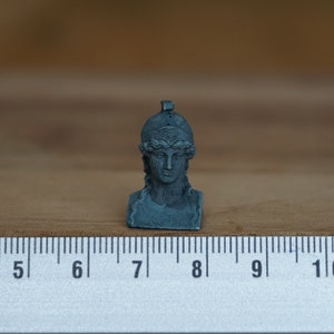May include: A small, gray, sculpted bust of a woman wearing a helmet. The bust is about 7 cm tall and is sitting on a ruler.