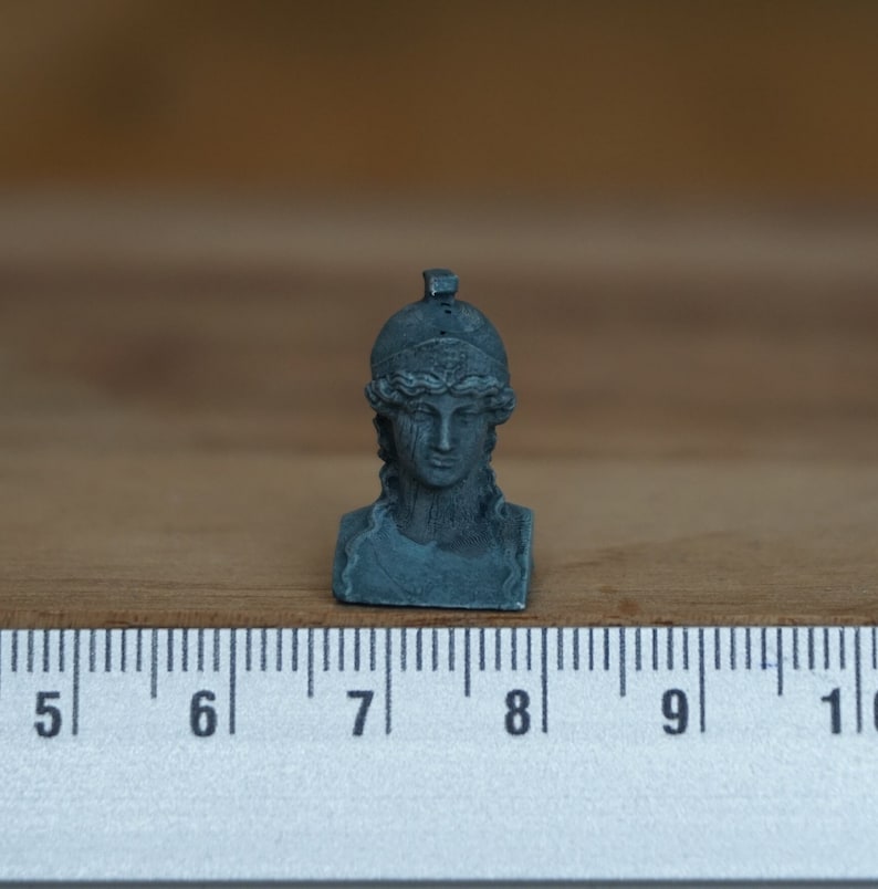 May include: A small, gray, sculpted bust of a woman wearing a helmet. The bust is about 7 cm tall and is sitting on a ruler.