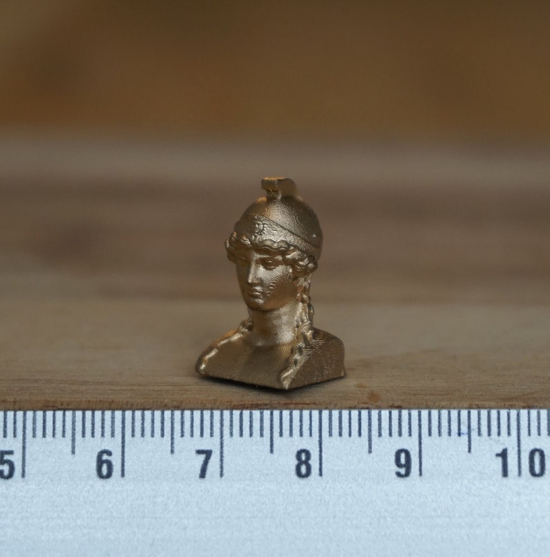 May include: A gold-colored bust of a woman wearing a helmet. The bust is about 2 inches tall and is sitting on a wooden surface.