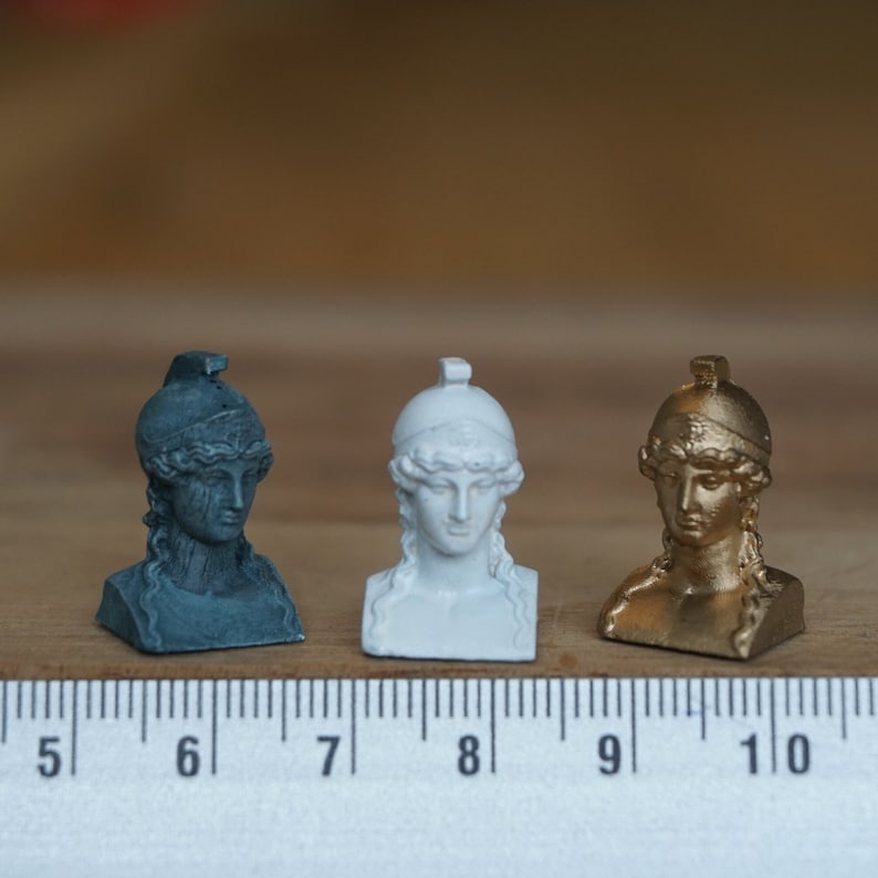May include: Three miniature busts of a woman wearing a helmet, each painted a different color: black, white, and gold. The busts are about 2 inches tall and are lined up on a ruler.