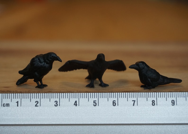 May include: Three black crow figurines on a wooden surface with a ruler for scale. The crows are approximately 2 inches tall.